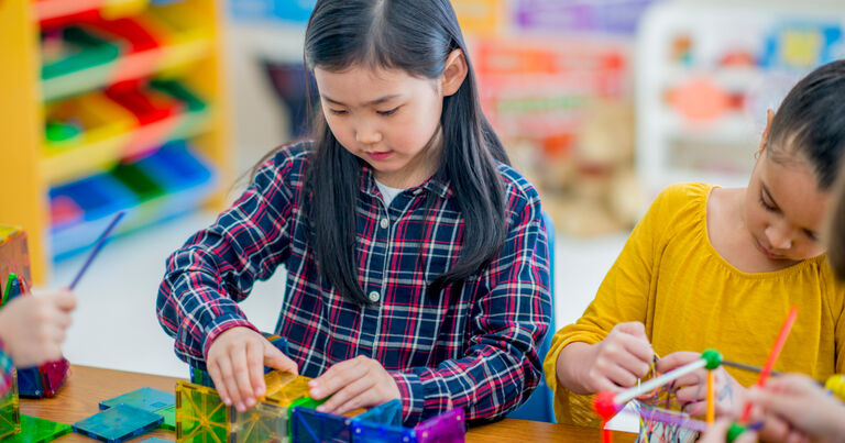 A young girl sits at a desk and plays with blocks. She sits next to a girl in a yellow shirt who is drawing with markers.